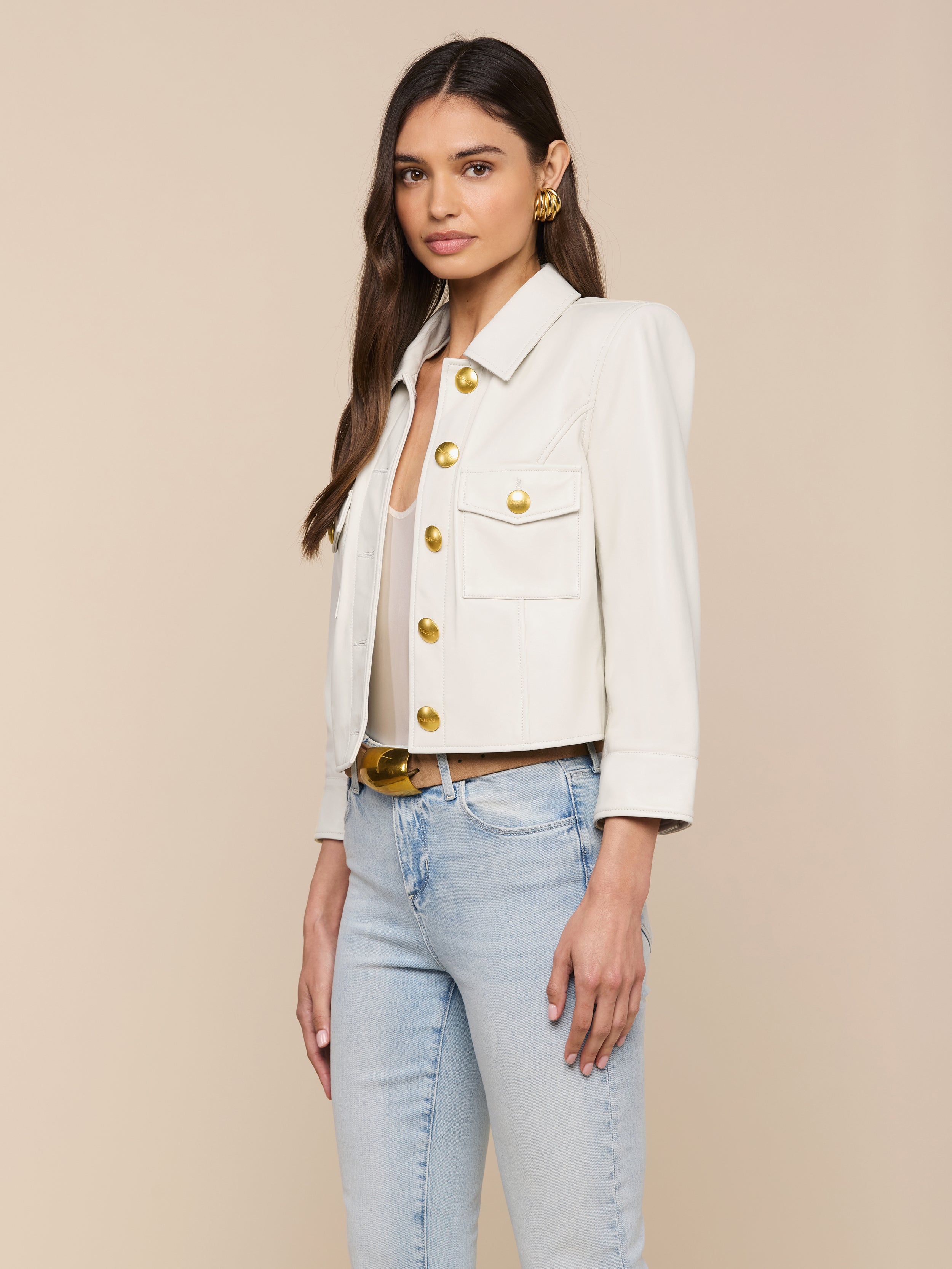 A woman wearing the Kumi Cropped Leather Jacket in white with large gold buttons, paired with light blue jeans and gold earrings, stands against an off-white background.