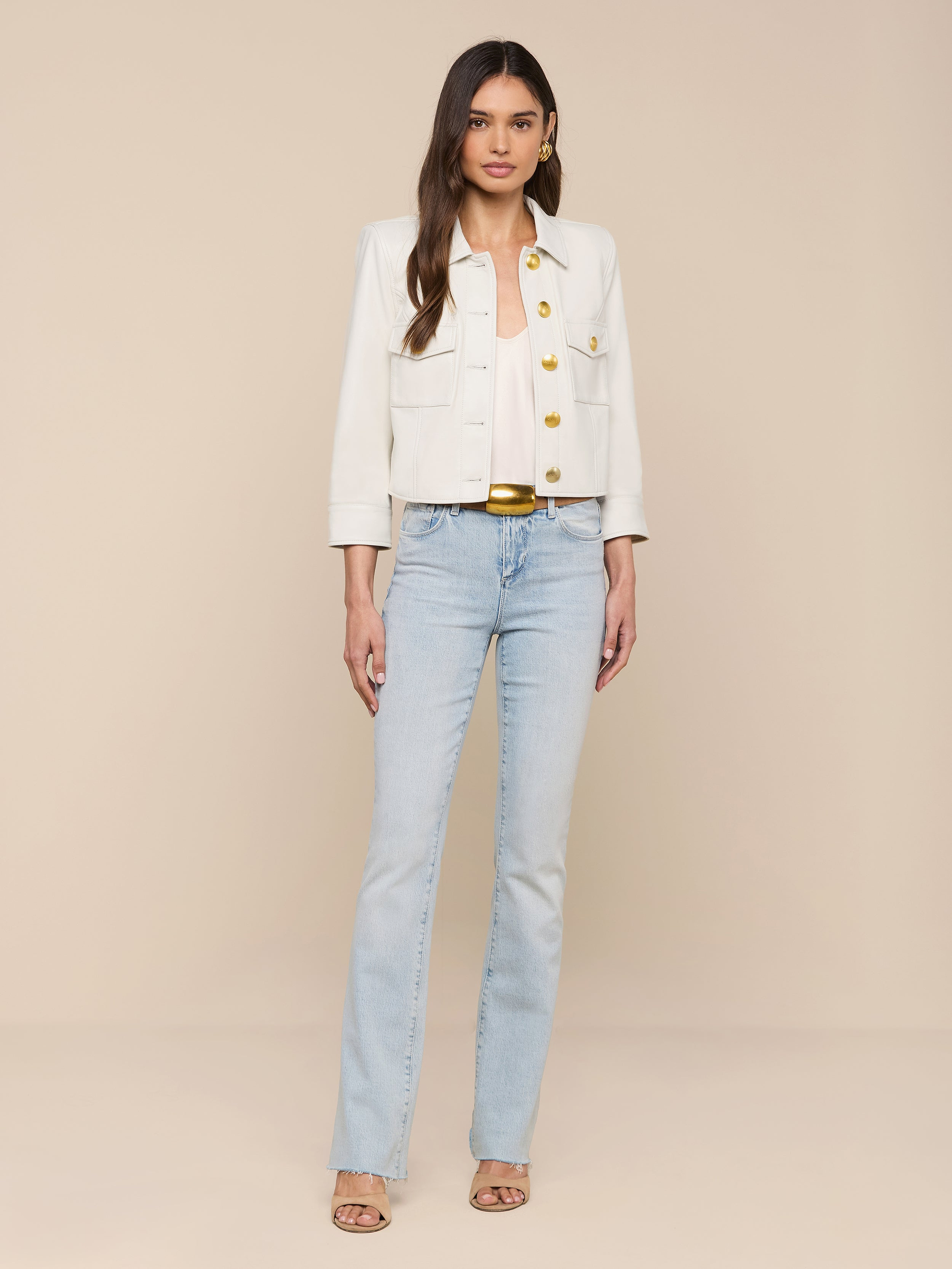A woman stands against a plain white background, wearing the Kumi Cropped Leather Jacket with gold buttons, a white top, light blue jeans, and beige heeled sandals. She has long brown hair and a neutral expression.