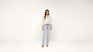 A woman stands facing forward against a plain white background, wearing a light beige buttoned jacket, light blue jeans, and beige heeled sandals.