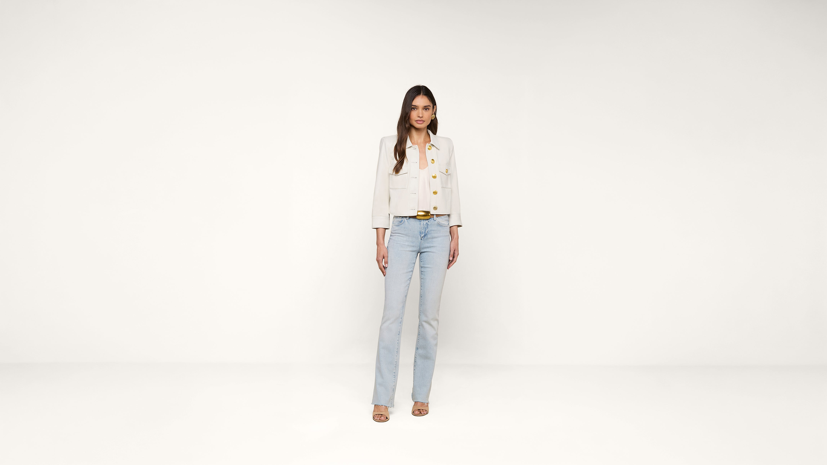 A woman stands facing forward against a plain white background, wearing a light beige buttoned jacket, light blue jeans, and beige heeled sandals.