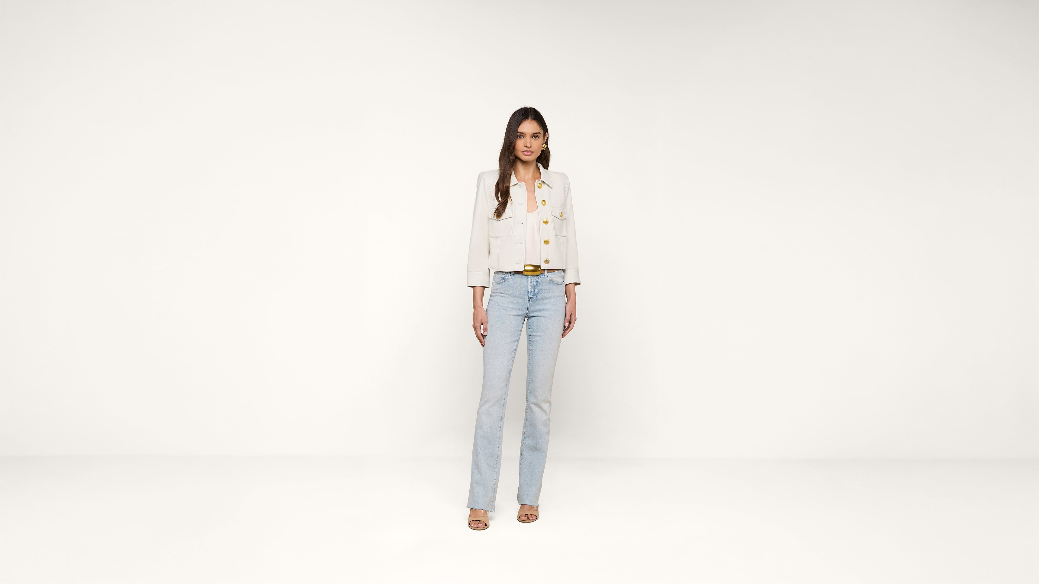 A woman stands facing forward against a plain white background, wearing a light beige buttoned jacket, light blue jeans, and beige heeled sandals.
