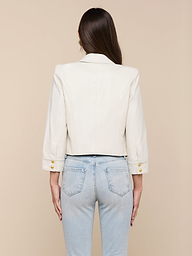A woman with long brown hair, wearing the Kumi Cropped Leather Jacket in white lambskin with gold buttons and light blue jeans, stands facing away against a plain background.