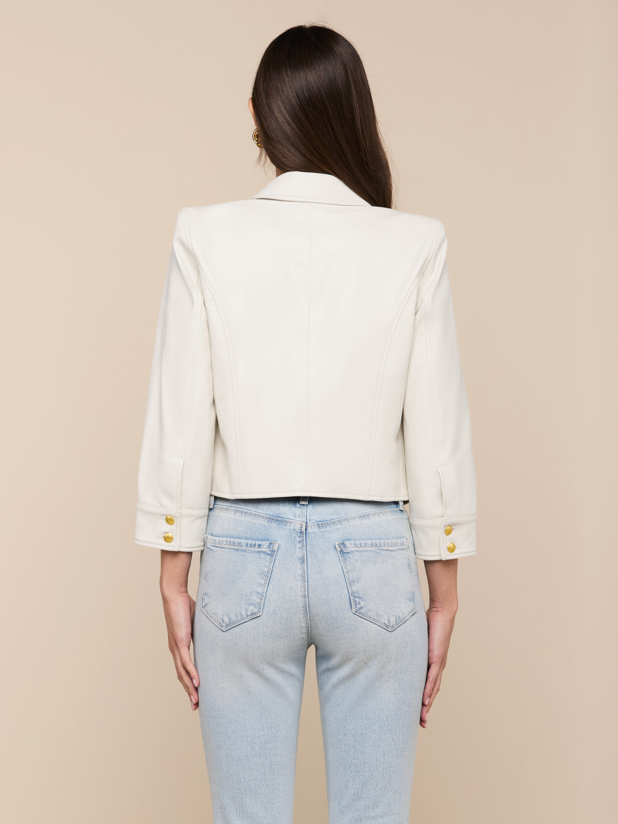 A woman with long brown hair, wearing the Kumi Cropped Leather Jacket in white lambskin with gold buttons and light blue jeans, stands facing away against a plain background.