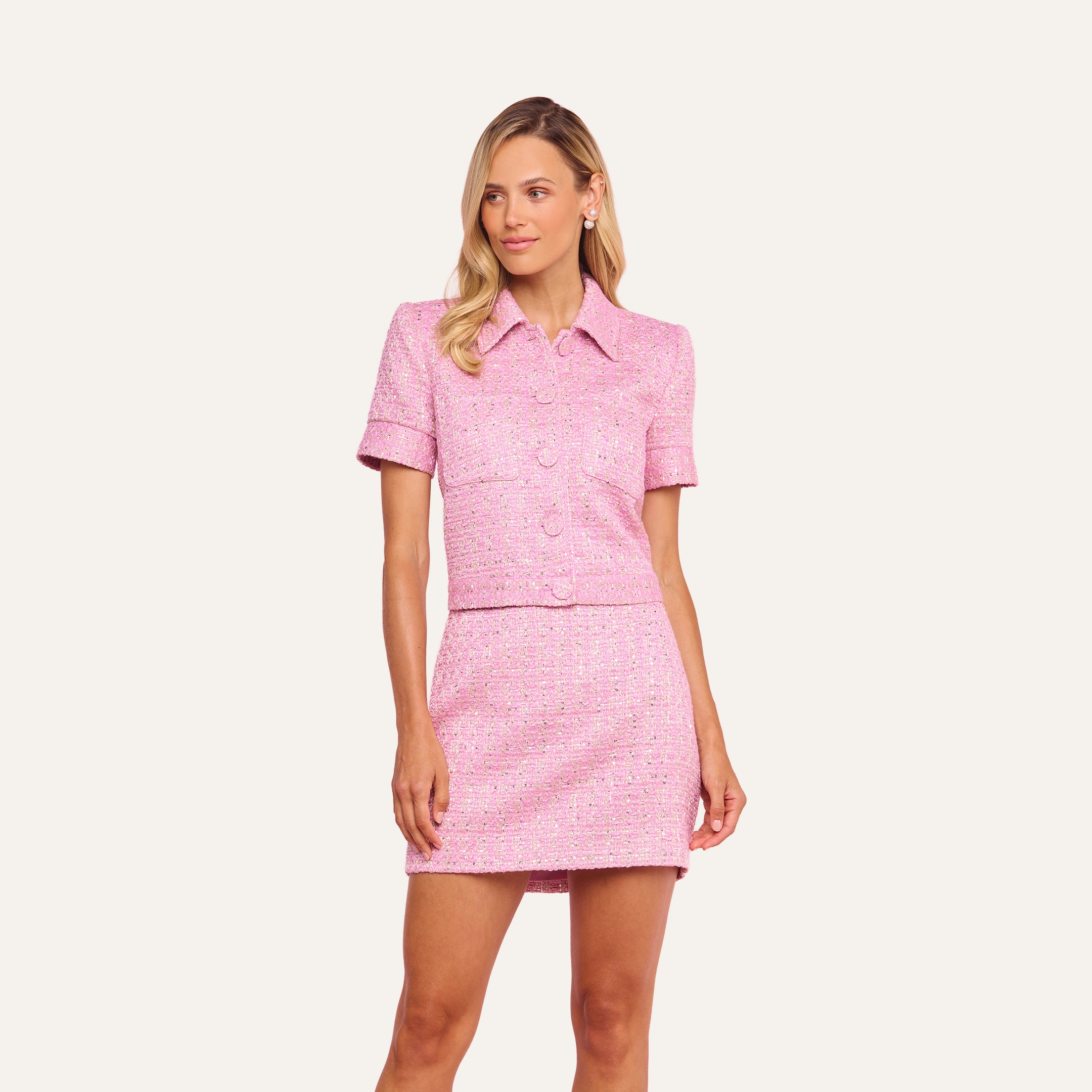 A woman wearing the Chita Tweed Jacket—a pink cropped tweed jacket with short sleeves—paired with a matching mini skirt, standing against a plain light background.