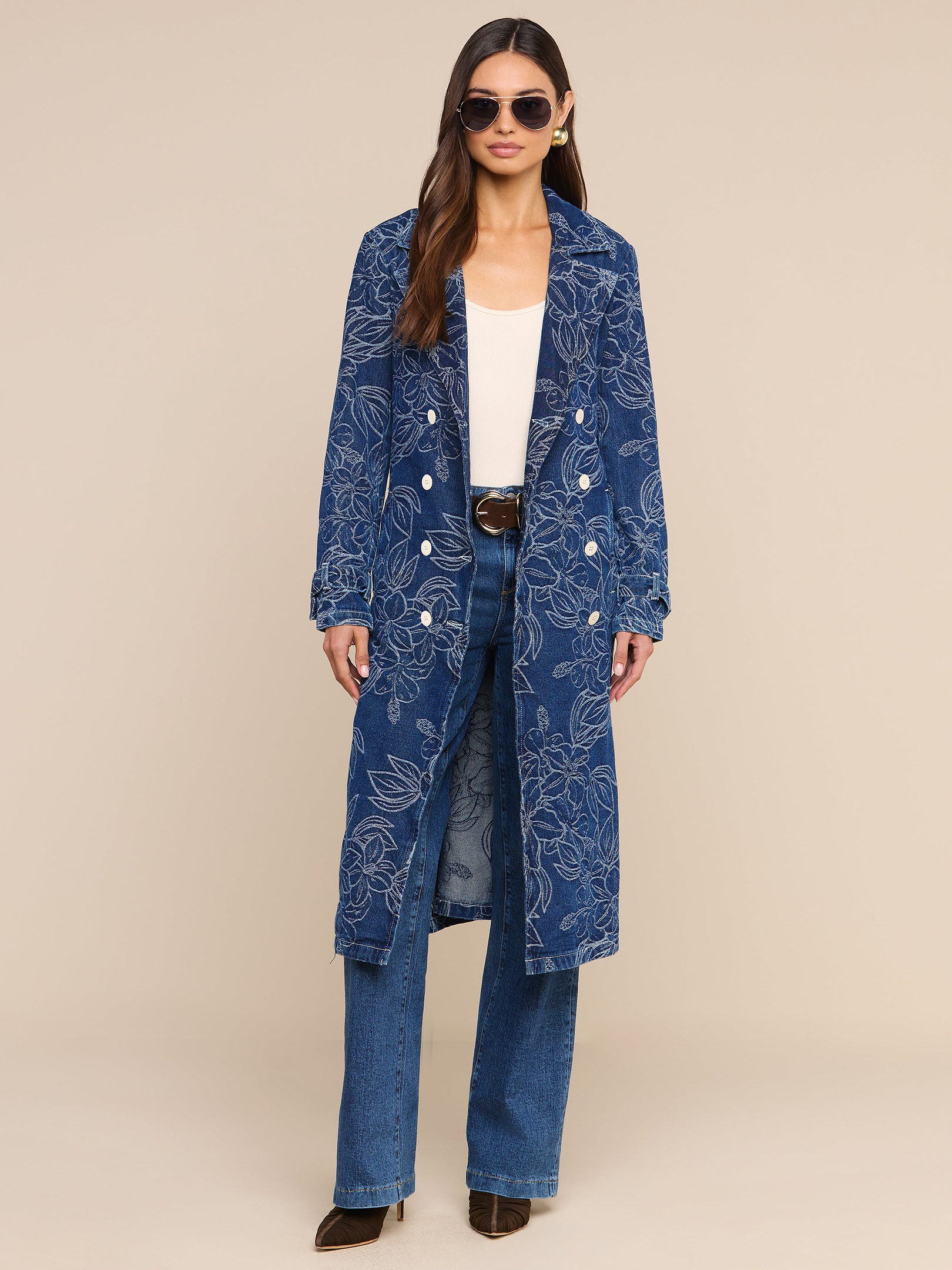 A woman wearing sunglasses and the Bradley Trench Coat—a long blue floral jacquard style—over a white top, blue jeans, and dark shoes stands against a plain white background.