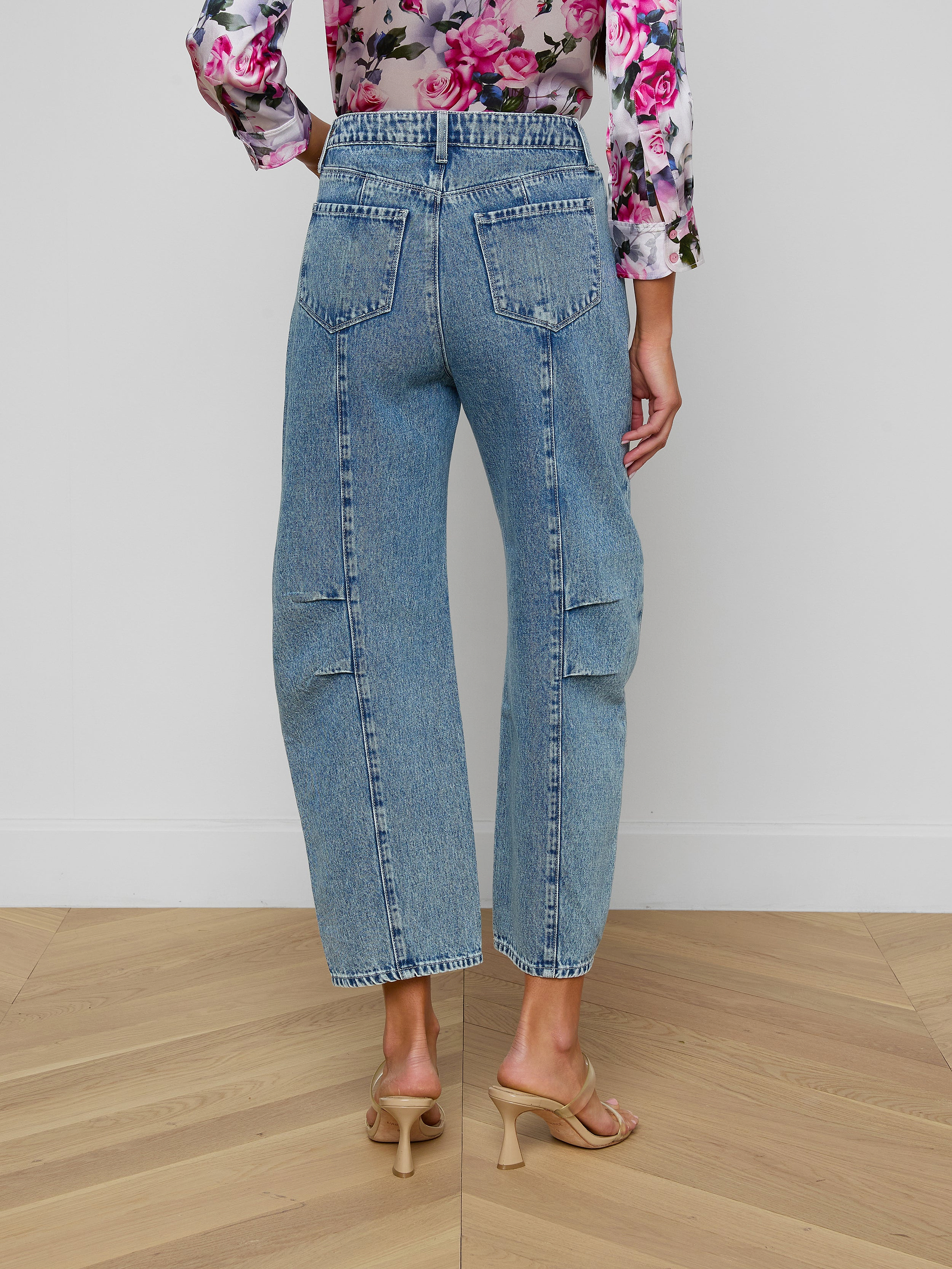 A person is seen from the back wearing the Reba Rodeo Jean in a barrel-leg silhouette with high rise, paired with beige heels. The long-sleeved top features a pink floral pattern, set against a plain light-colored background.