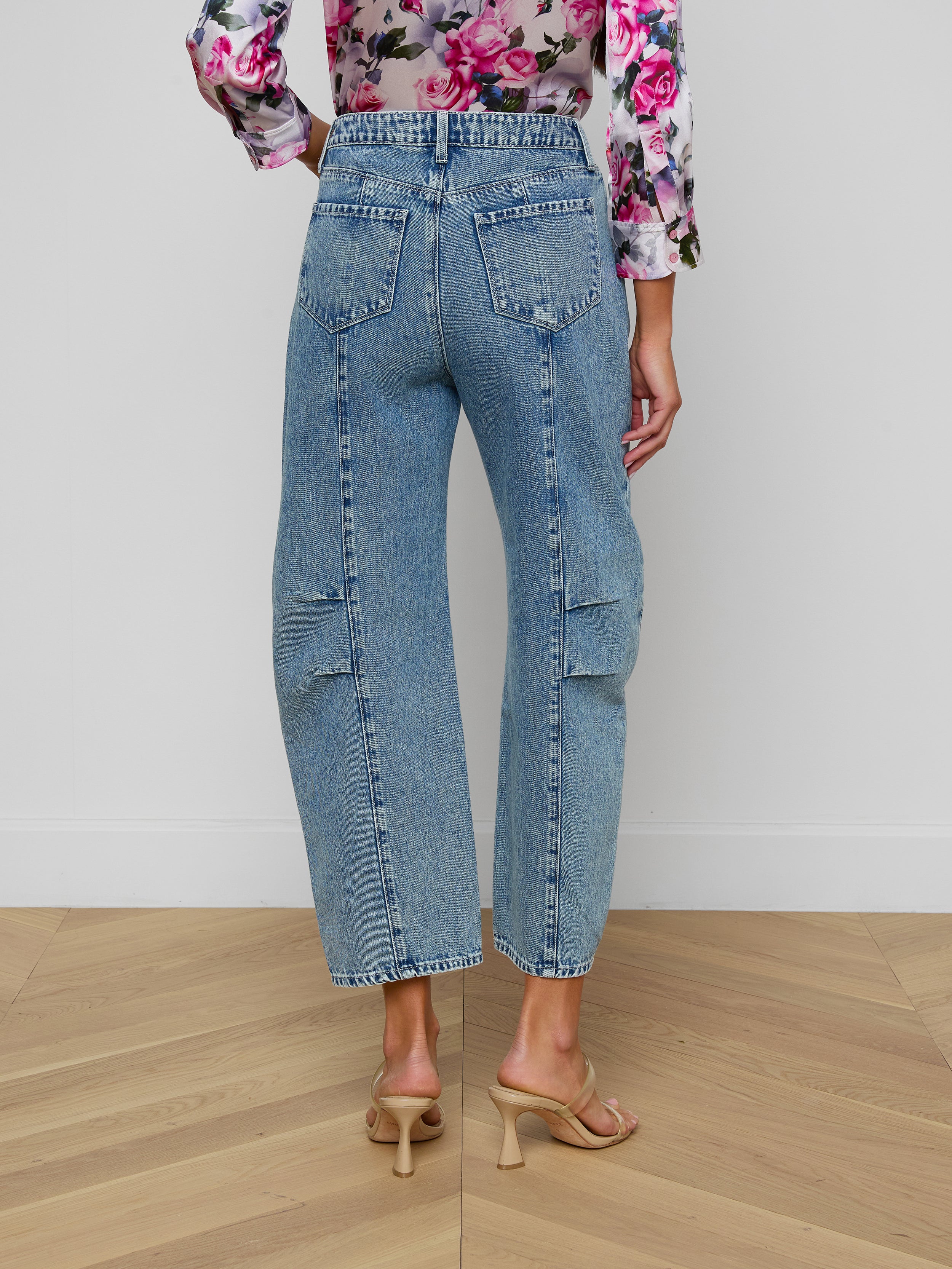 A person is seen from the back wearing the Reba Rodeo Jean in a barrel-leg silhouette with high rise, paired with beige heels. The long-sleeved top features a pink floral pattern, set against a plain light-colored background.