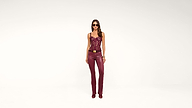 A woman stands against a white background, wearing sunglasses, a maroon lace top, maroon leather pants with a gold belt, maroon shoes, and gold bracelets.
