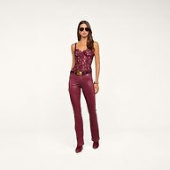 A woman wearing sunglasses, a burgundy lace corset top, and the Selma Coated Bootcut Jean with a belt and matching shoes stands against a plain white background.