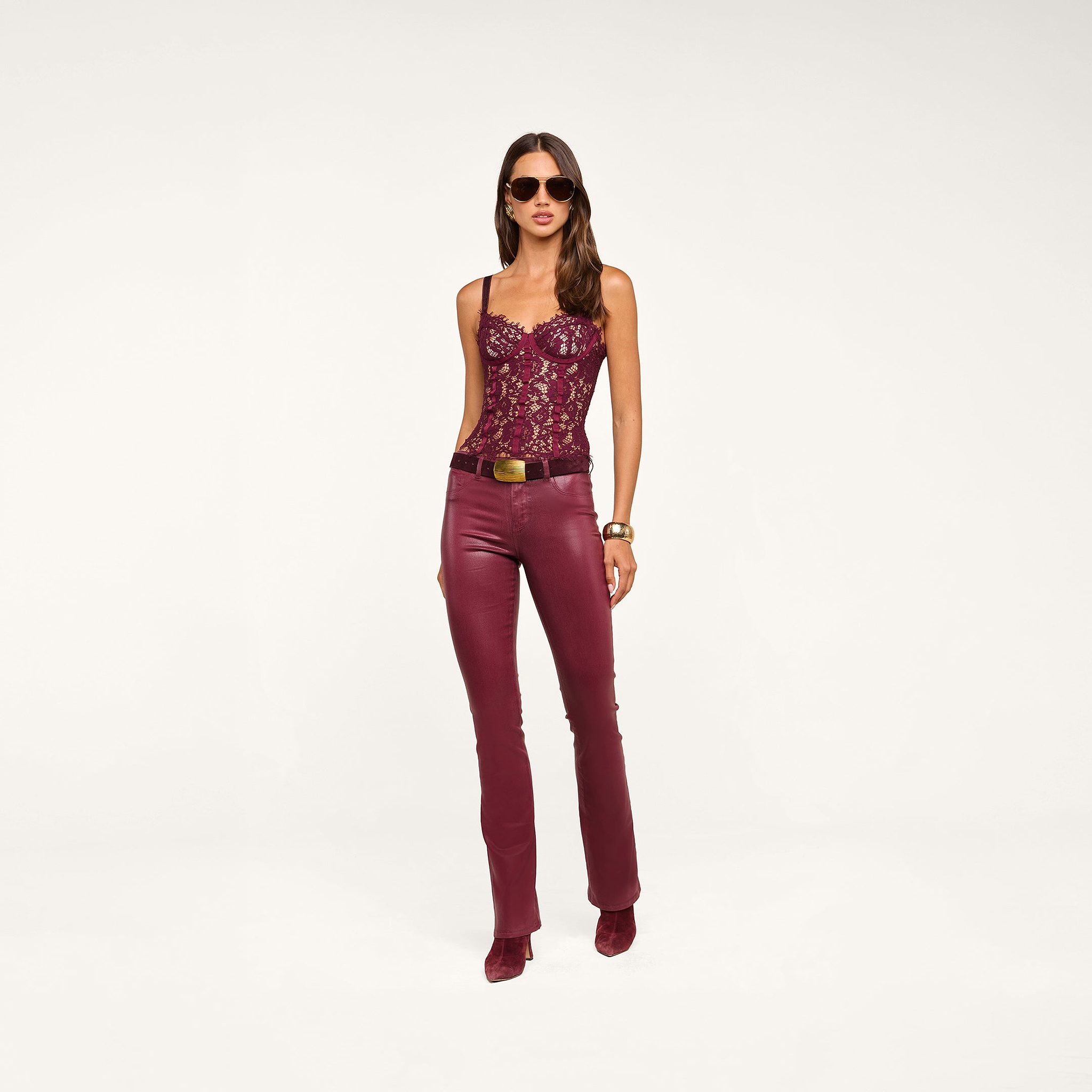 A woman wearing sunglasses, a burgundy lace corset top, and the Selma Coated Bootcut Jean with a belt and matching shoes stands against a plain white background.
