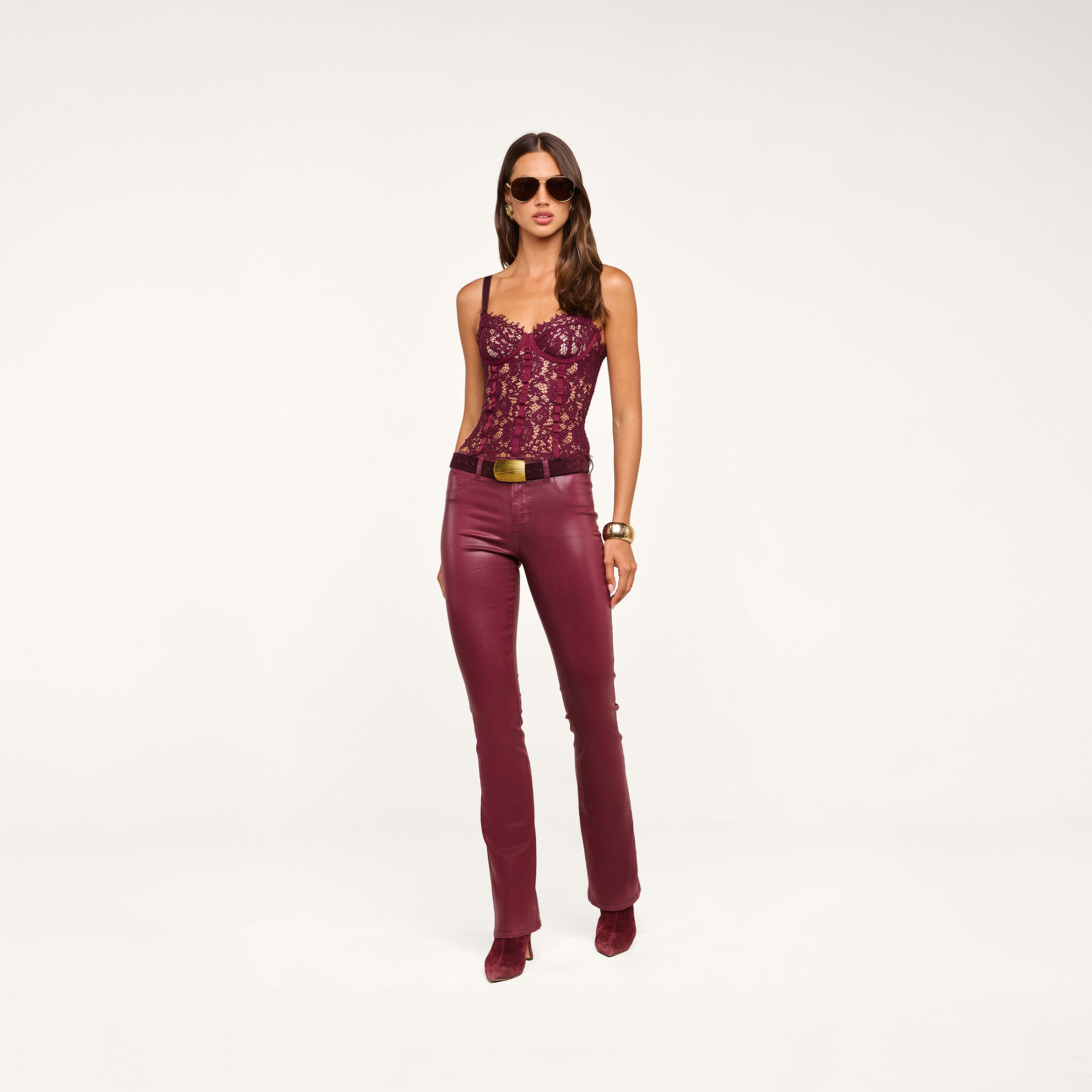 A woman wearing sunglasses, a burgundy lace corset top, and the Selma Coated Bootcut Jean with a belt and matching shoes stands against a plain white background.