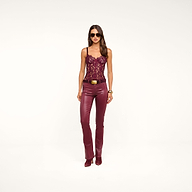 A woman wearing sunglasses, a burgundy lace top, the Selma Coated Bootcut Jean in maroon leather-alternative denim, and boots stands against a plain white background.