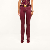 A woman is standing with her arms by her sides against a plain background, wearing high-rise Selma Coated Bootcut Jeans paired with a maroon leather-alternative lace top, matching boots, a black belt featuring a gold buckle, and a gold bracelet.