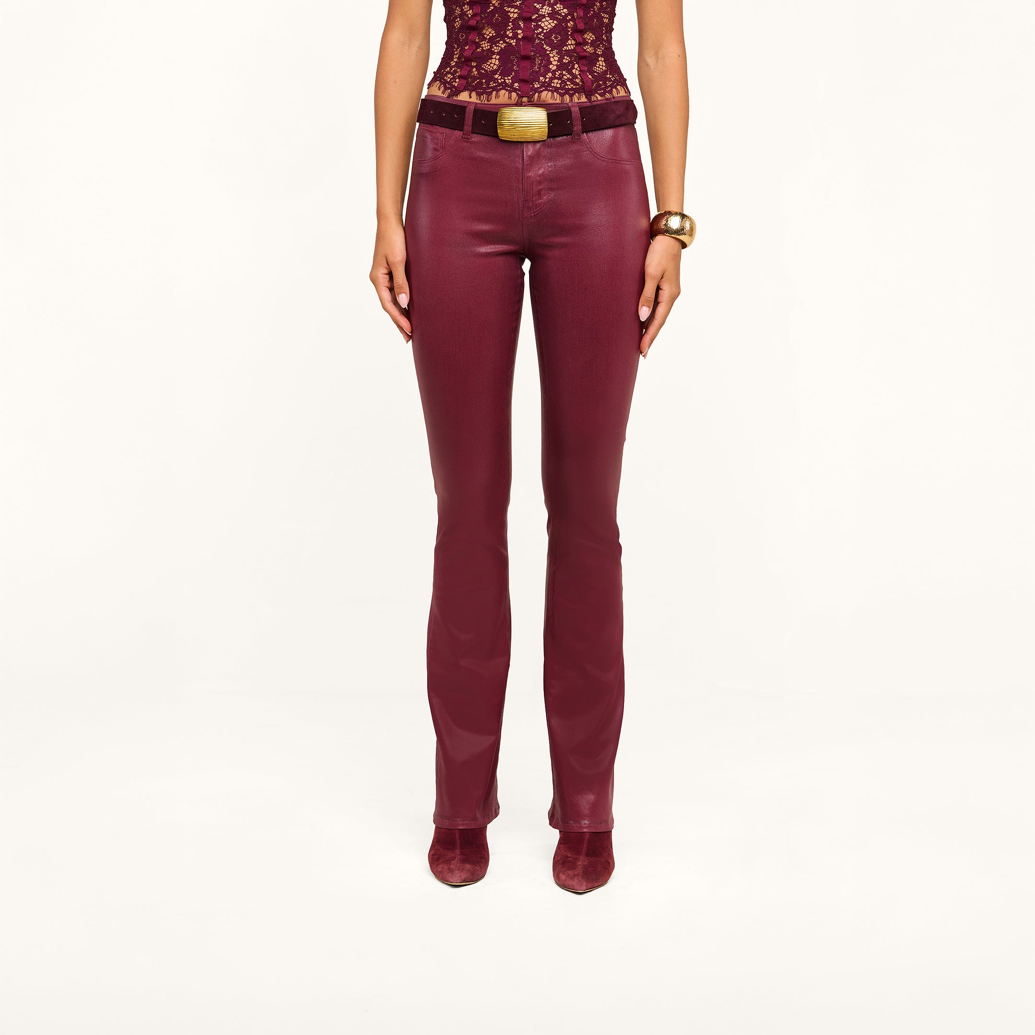 A woman is standing with her arms by her sides against a plain background, wearing high-rise Selma Coated Bootcut Jeans paired with a maroon leather-alternative lace top, matching boots, a black belt featuring a gold buckle, and a gold bracelet.