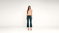 Woman wearing sunglasses, a sleeveless beige top, teal flared pants, and sandals stands against a plain white background, facing forward with a neutral expression.