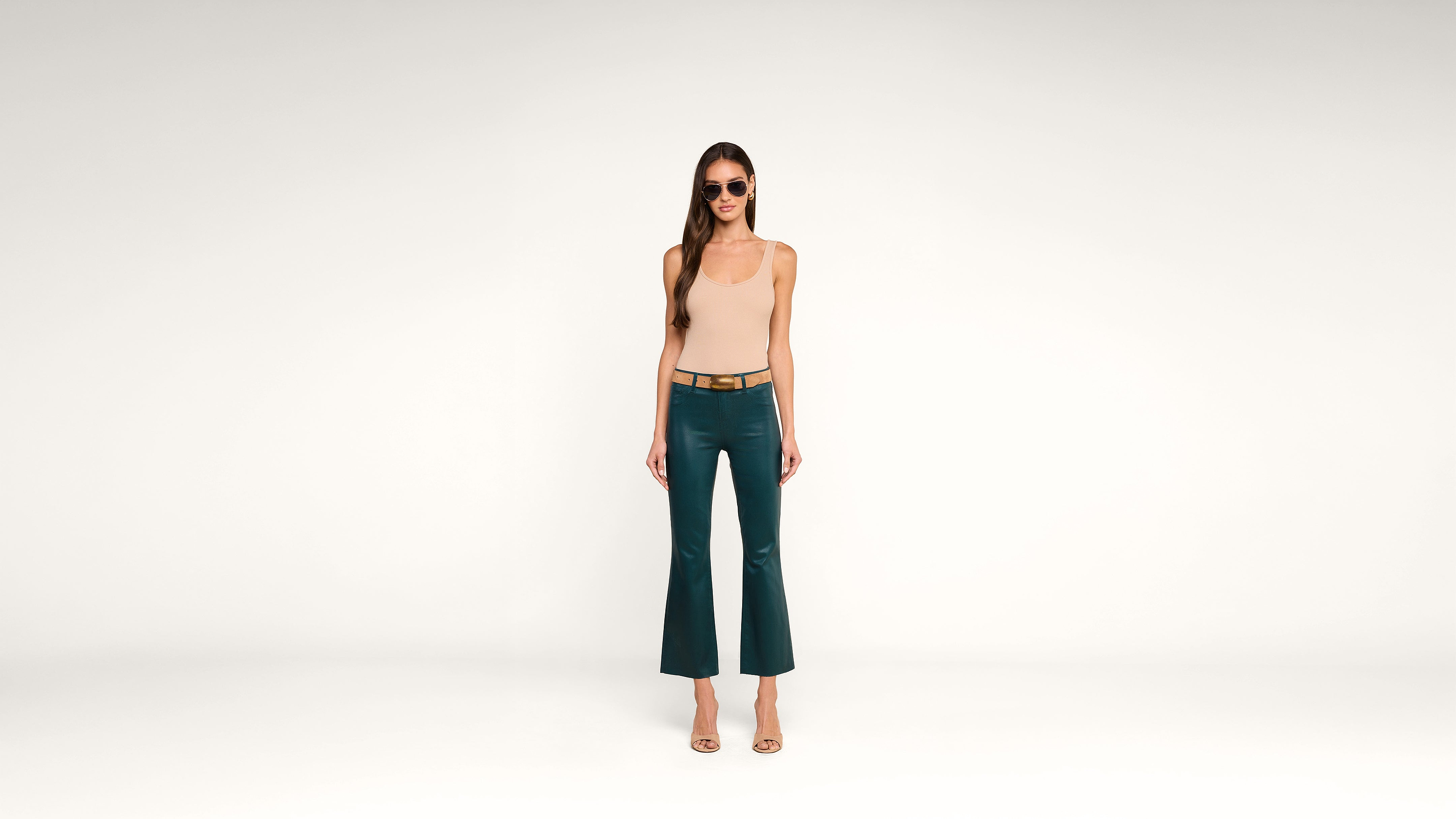 Woman wearing sunglasses, a sleeveless beige top, teal flared pants, and sandals stands against a plain white background, facing forward with a neutral expression.