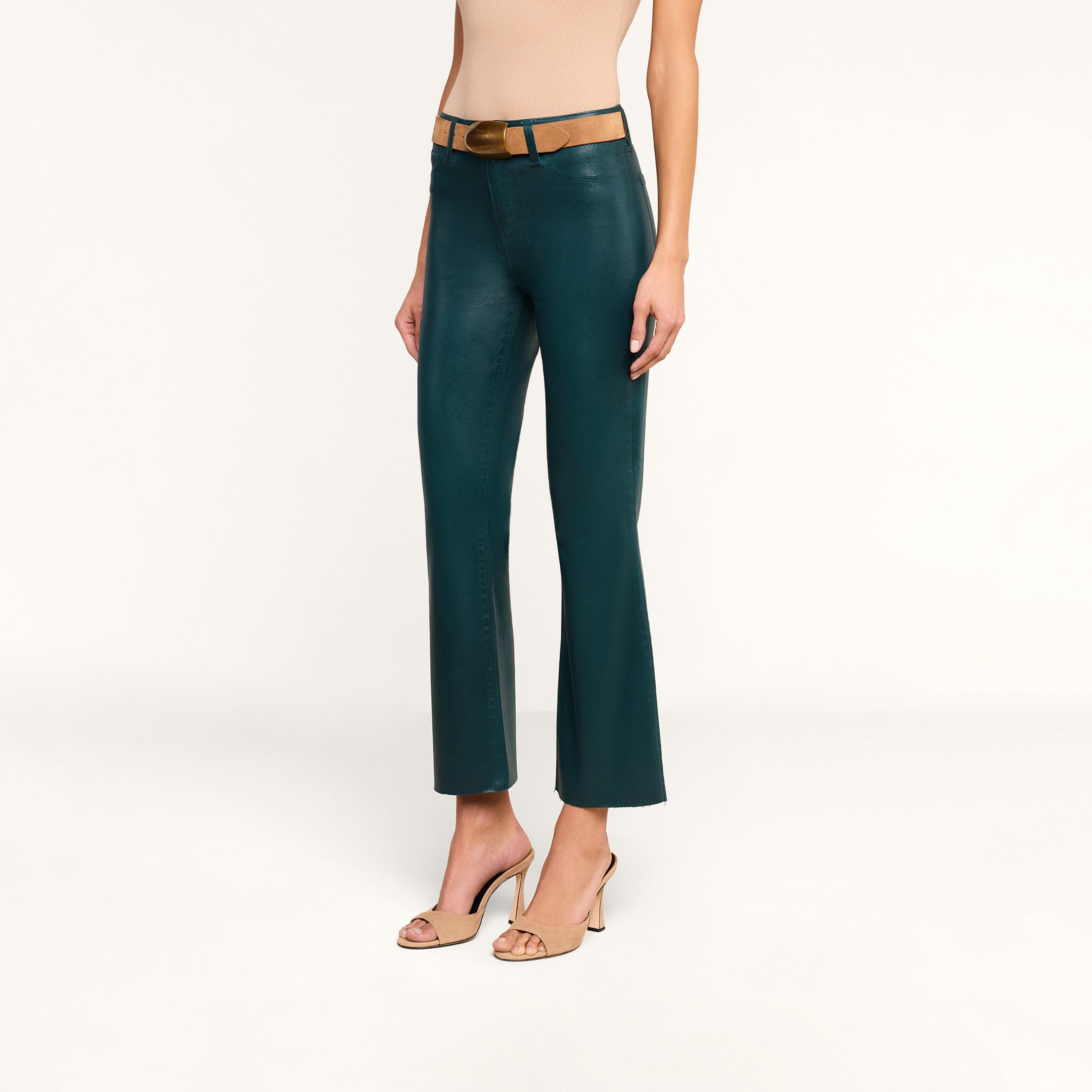 A woman wearing the Kendra Coated Cropped Flare Jean in teal, paired with a tan belt, a sleeveless beige top, and nude high-heeled sandals stands against a white background.