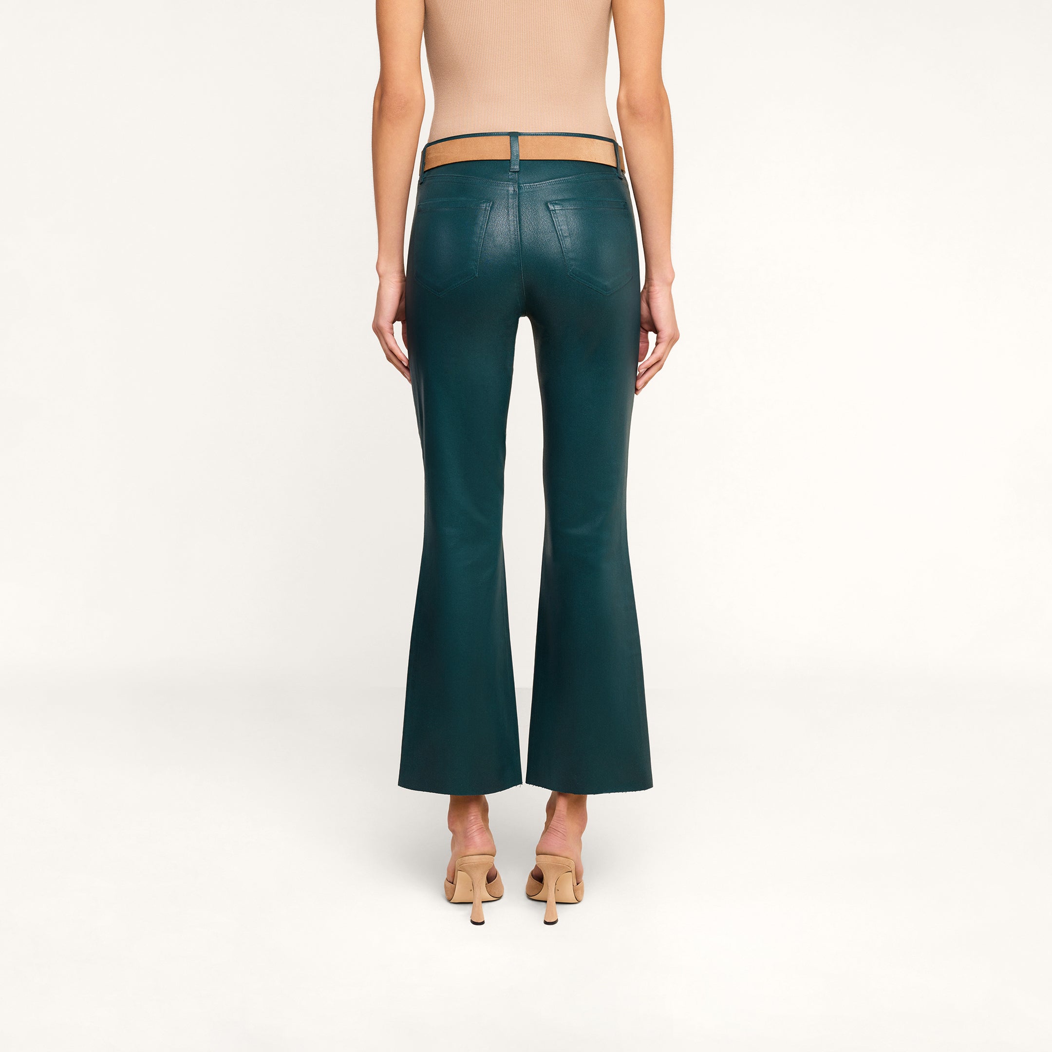 A person stands facing away, wearing the Kendra Coated Cropped Flare Jean in dark teal, paired with a tan sleeveless top and tan high-heeled shoes against a plain white background.