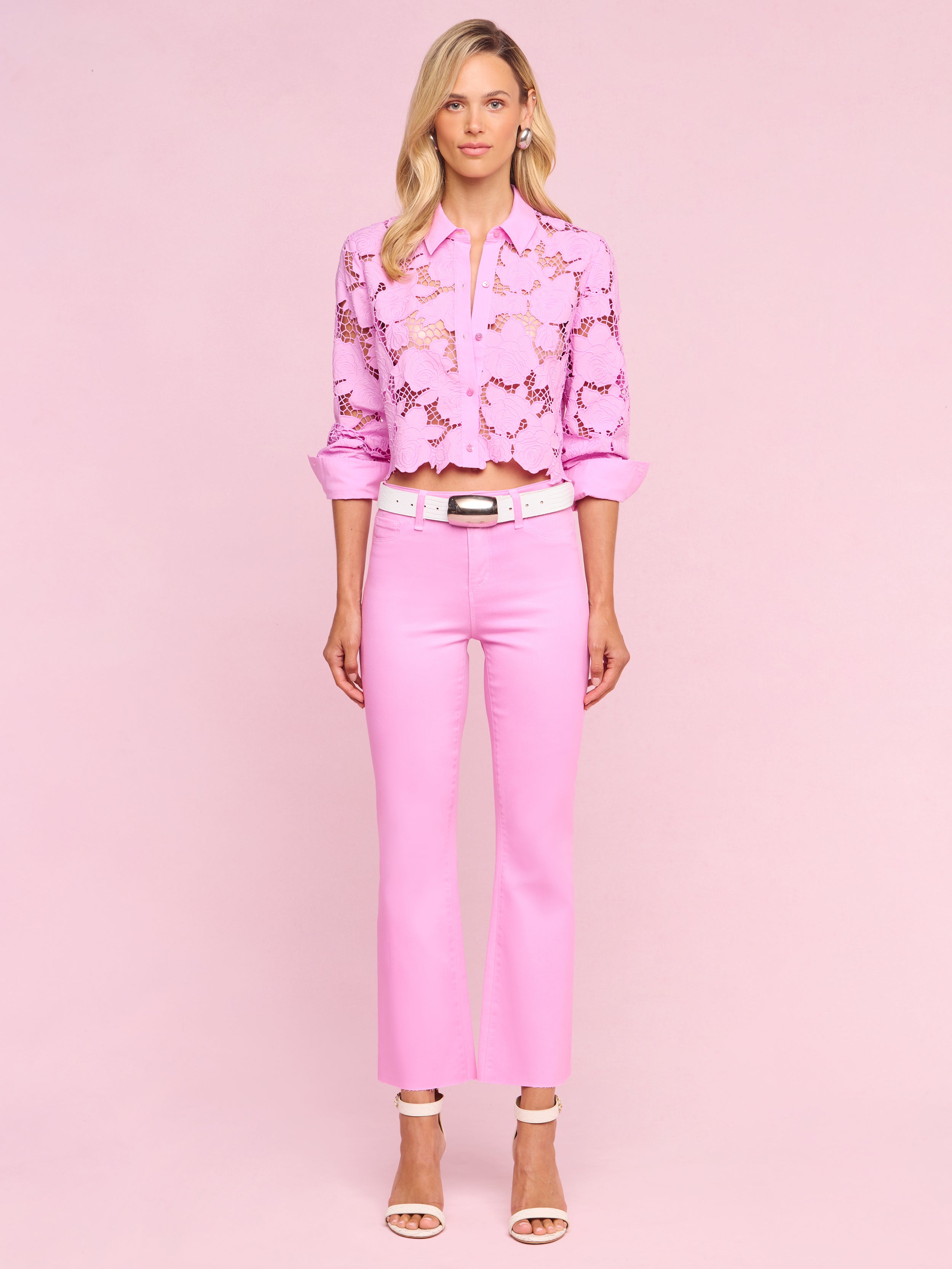 A woman with blonde hair and a neutral expression stands against a white background wearing the Kendra Coated Cropped Flare Jean in pink, paired with a light pink floral blouse, a white belt, and white heeled sandals.