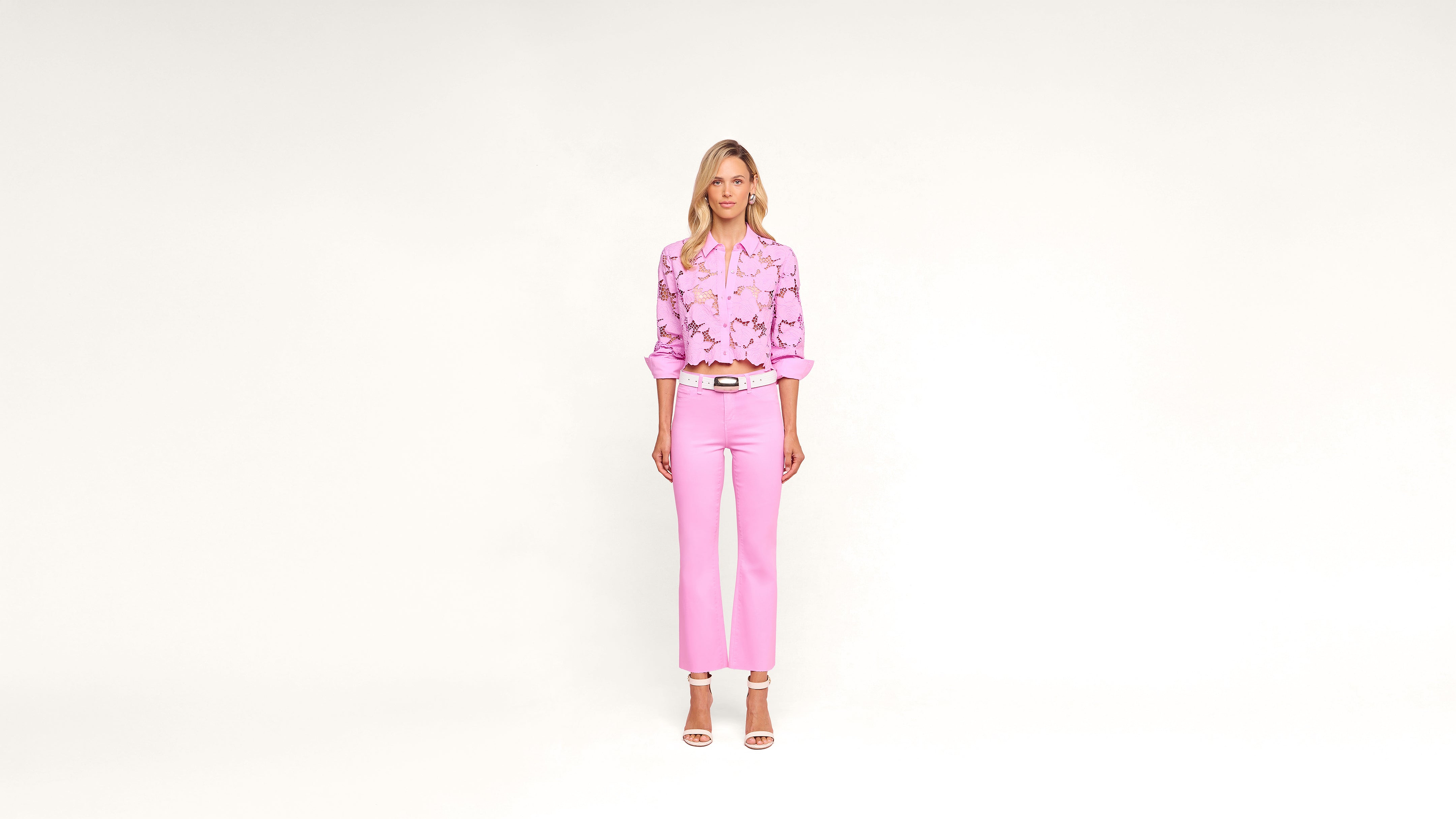 A woman stands against a plain white background, wearing a pink floral blouse, light pink pants, white belt, and white heeled sandals. She faces the camera with a neutral expression.