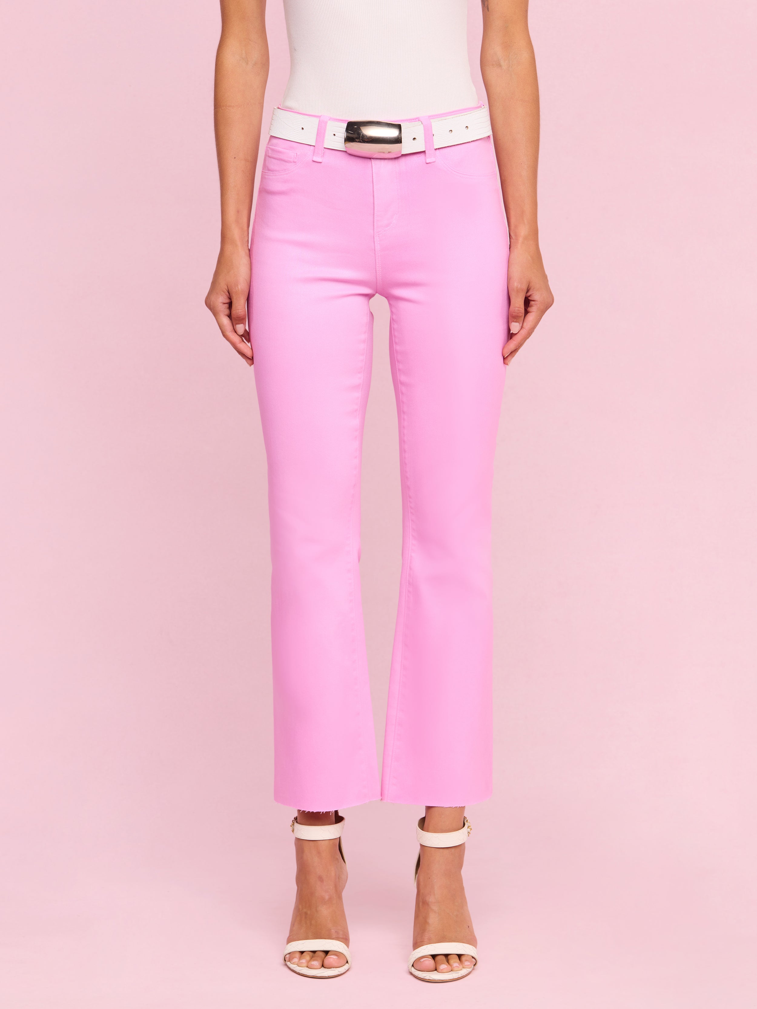 Wearing the Kendra Coated Cropped Flare Jean in bright pink coated denim, styled with a white belt and white high-heeled sandals, a person stands against a plain light background.