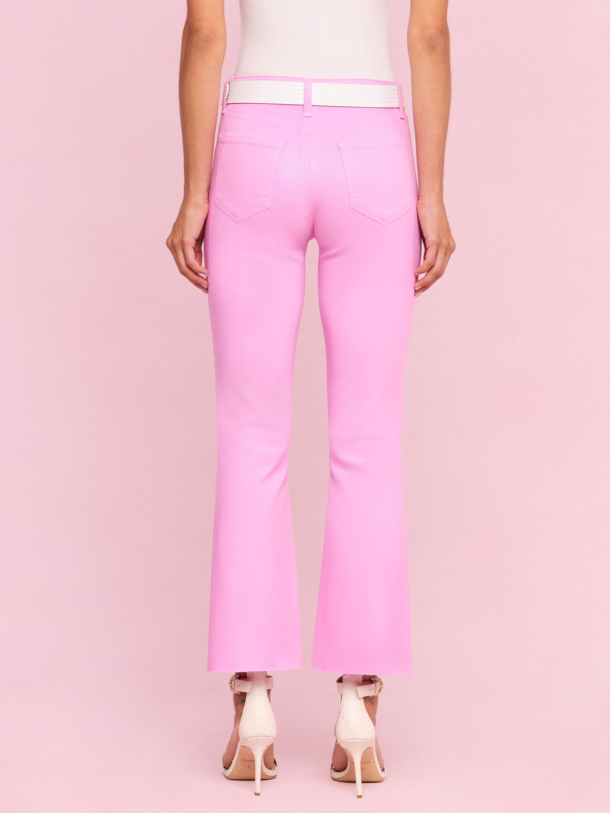 A person stands facing away, wearing the Kendra Coated Cropped Flare Jean in light pink crafted from coated denim, paired with high-heeled pink shoes against a plain, light background.