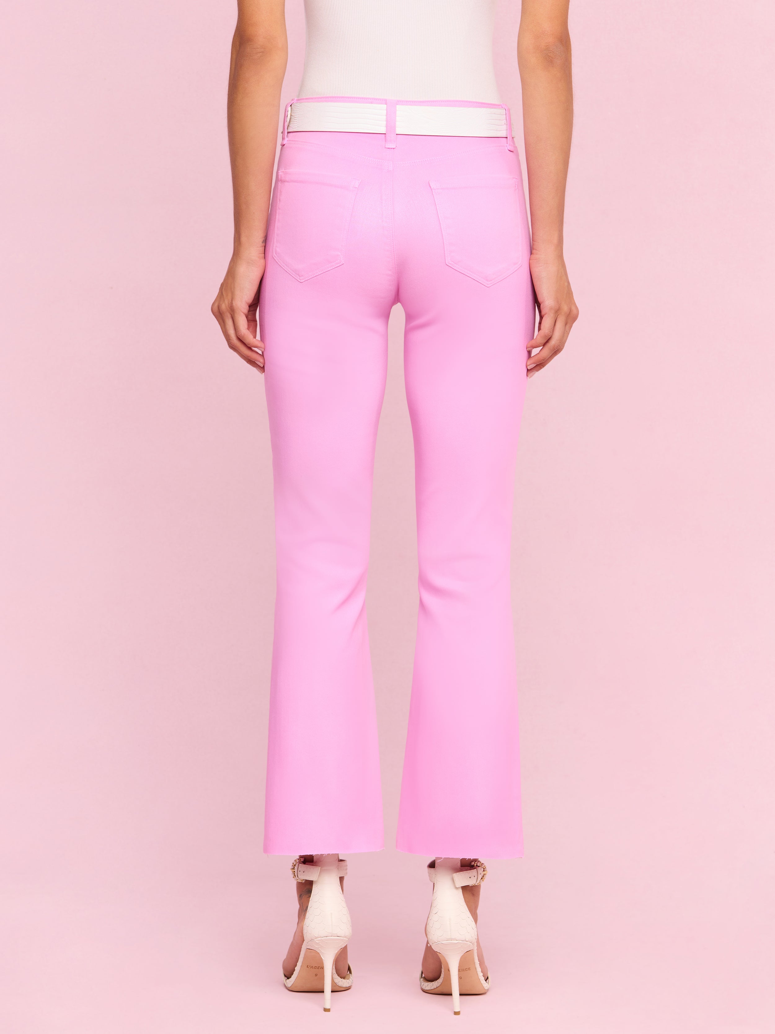 A person stands facing away, wearing the Kendra Coated Cropped Flare Jean in light pink crafted from coated denim, paired with high-heeled pink shoes against a plain, light background.