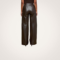 Indoors, a person is seen from the back wearing the high-waisted Livvy Leather Trouser by L’AGENCE in chocolate brown with a sleeveless top, standing on chevron-patterned wood flooring.