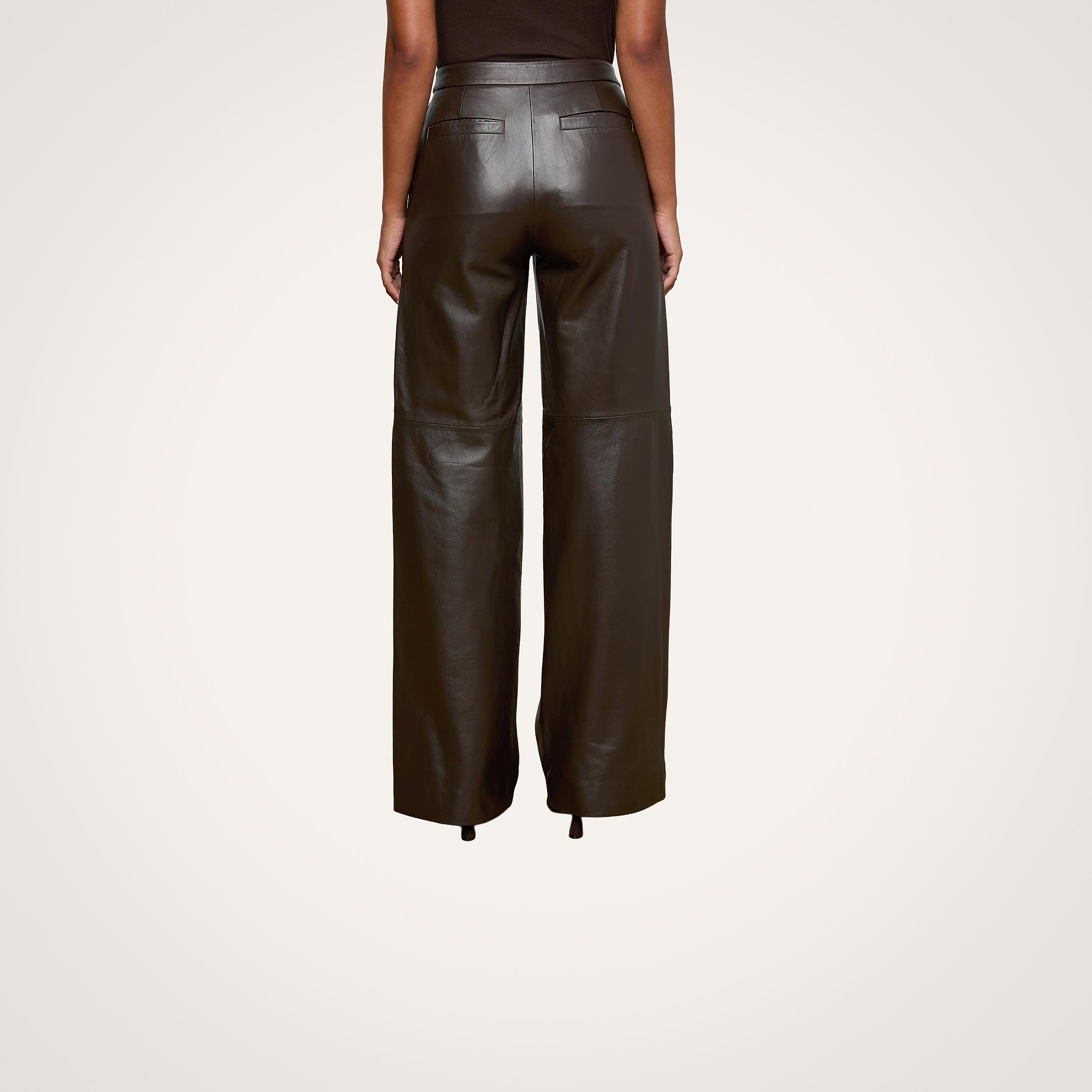 Indoors, a person is seen from the back wearing the high-waisted Livvy Leather Trouser by L’AGENCE in chocolate brown with a sleeveless top, standing on chevron-patterned wood flooring.