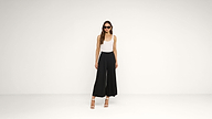 Woman wearing sunglasses, a sleeveless white top, black wide-leg pants, and white high-heeled sandals stands in a plain, light-colored room with a minimalist background.