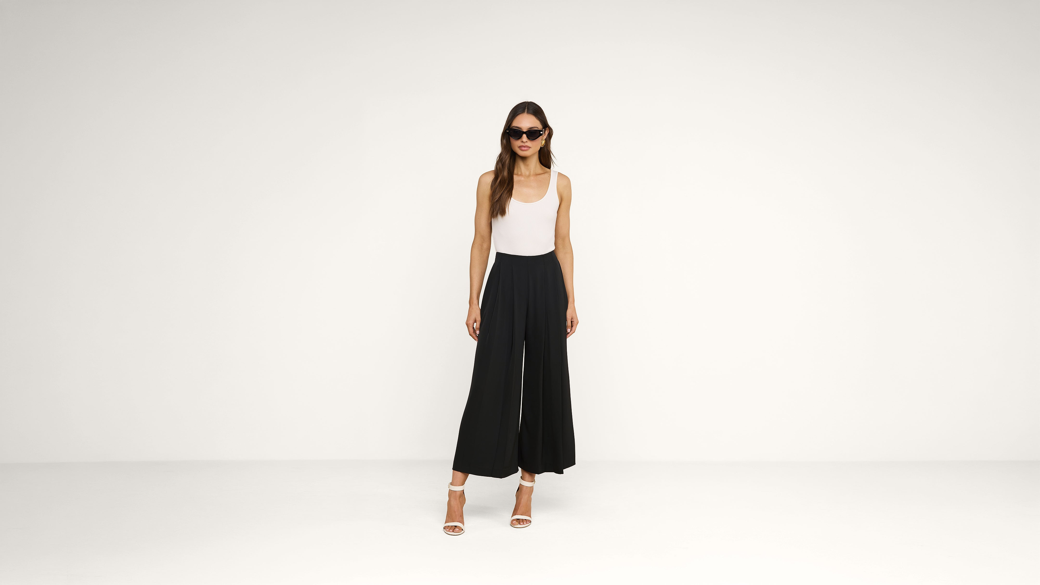 Woman wearing sunglasses, a sleeveless white top, black wide-leg pants, and white high-heeled sandals stands in a plain, light-colored room with a minimalist background.