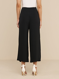 Wearing the Novalynn Cropped Pleated Pant, a person stands facing away in a minimal, white space, paired with a white sleeveless top and white high-heeled sandals.
