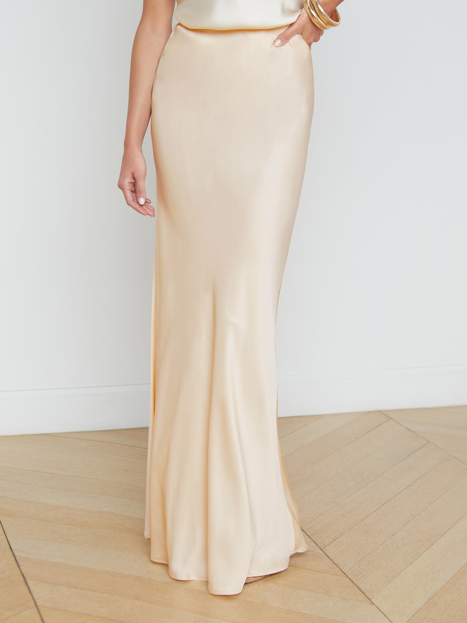 A person models the Zeta Satin Maxi Skirt—a long, cream-colored, high-waisted, bias-cut satin piece that gracefully flows to the floor—paired with gold bracelets and photographed against a plain white background. Only the lower half of the body is visible.