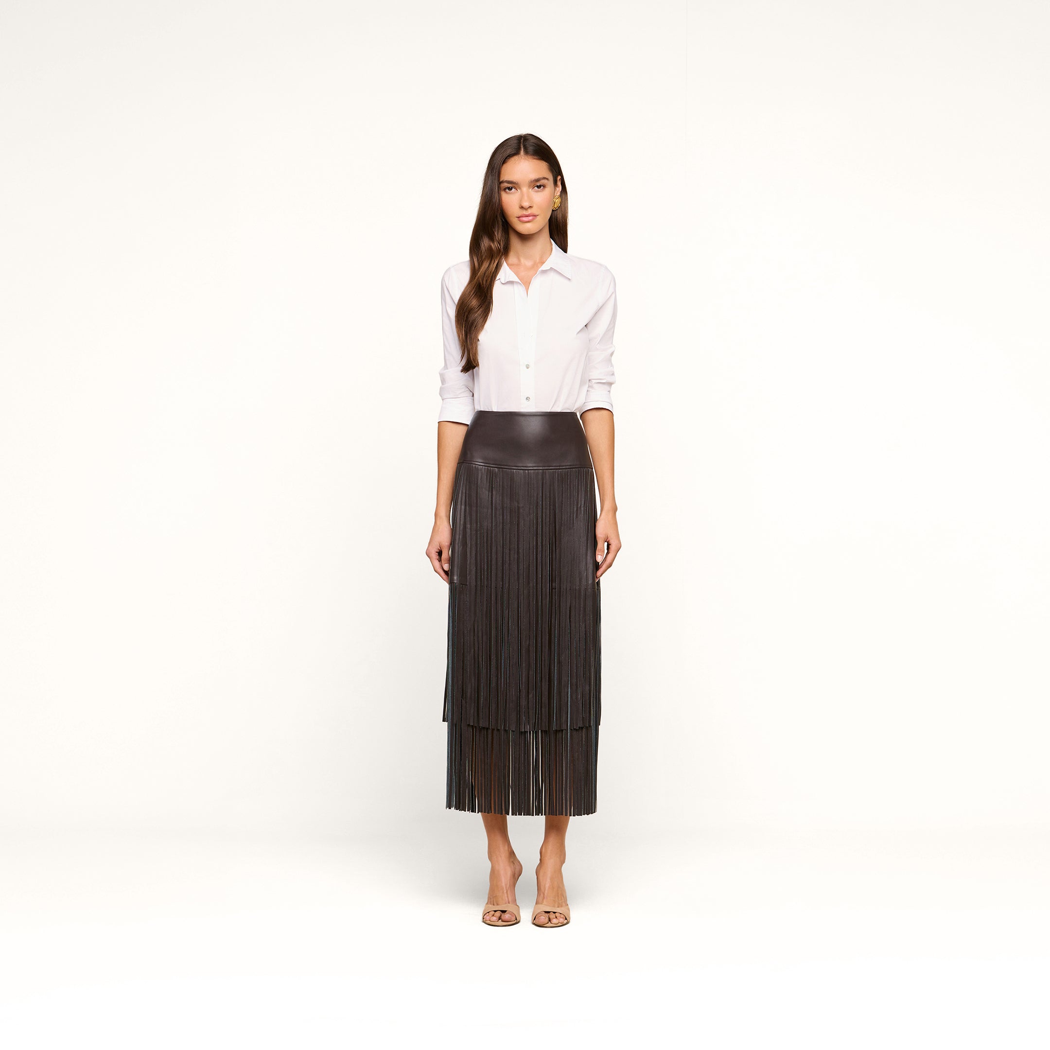 A woman stands facing forward against a plain white background, wearing a white button-down shirt, the Karolina Fringe Skirt in black, and beige open-toed heels.