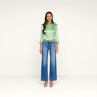 A woman stands against a plain white background, wearing the Dani Silk Blouse in light green, paired with blue wide-leg jeans, a tan belt, and beige heels. She has long brown hair and a neutral expression.