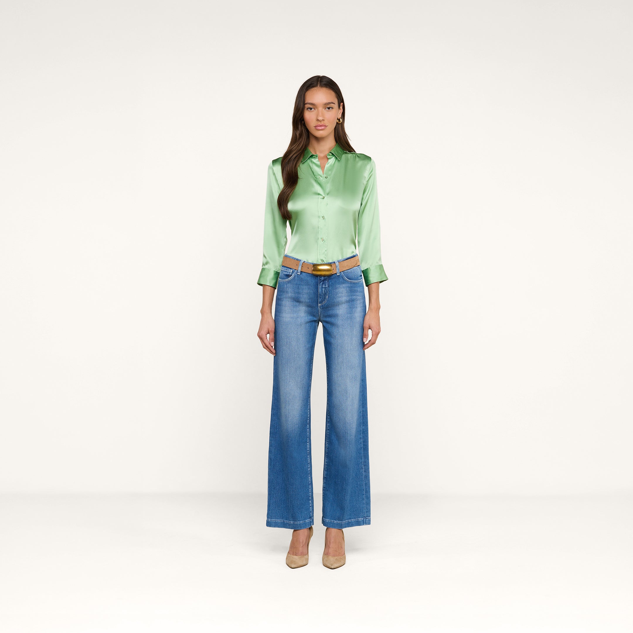 A woman stands against a plain white background, wearing the Dani Silk Blouse in light green, paired with blue wide-leg jeans, a tan belt, and beige heels. She has long brown hair and a neutral expression.