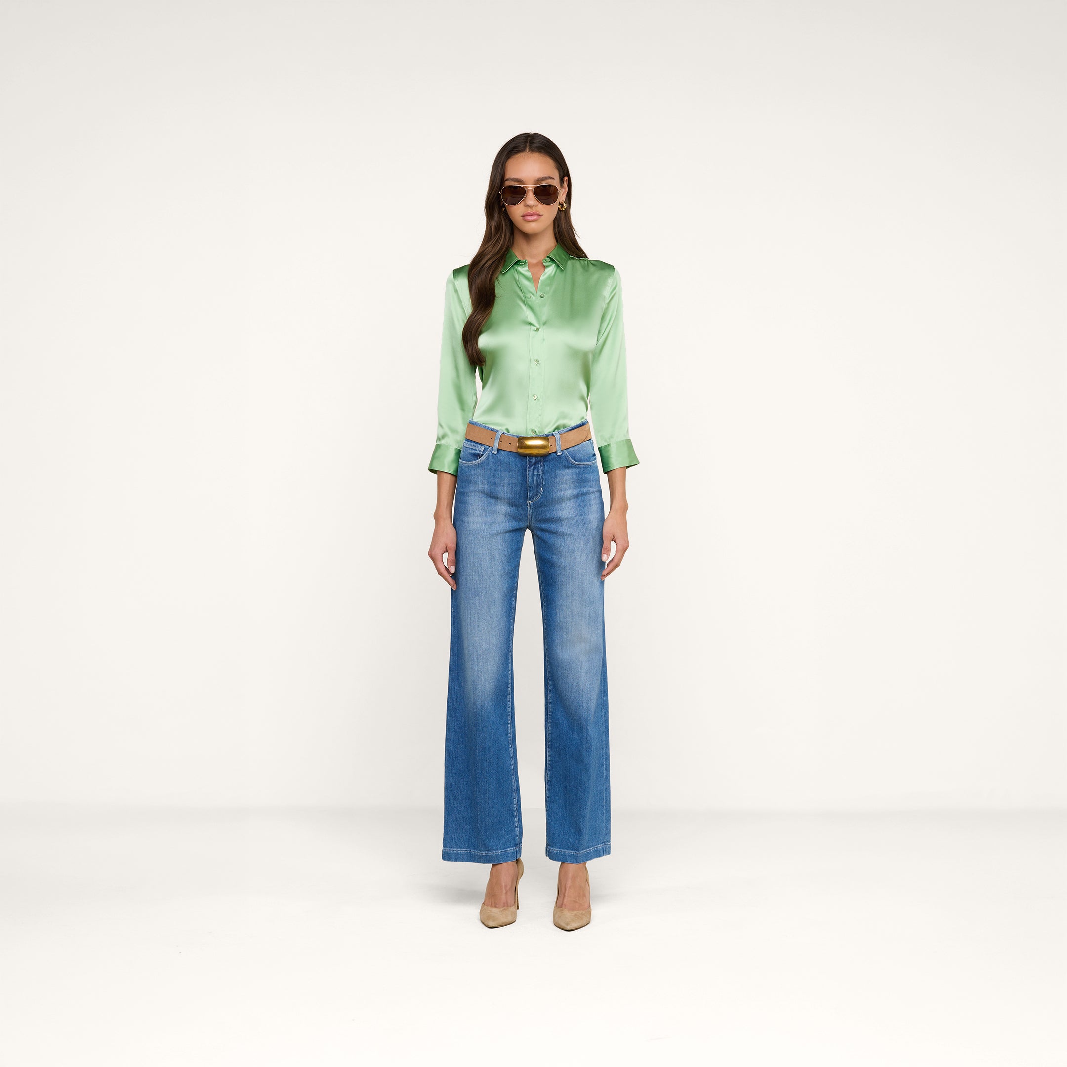 A woman stands against a white background wearing sunglasses, a brown belt, blue wide-leg jeans, beige heels, and the Dani Silk Blouse in green.