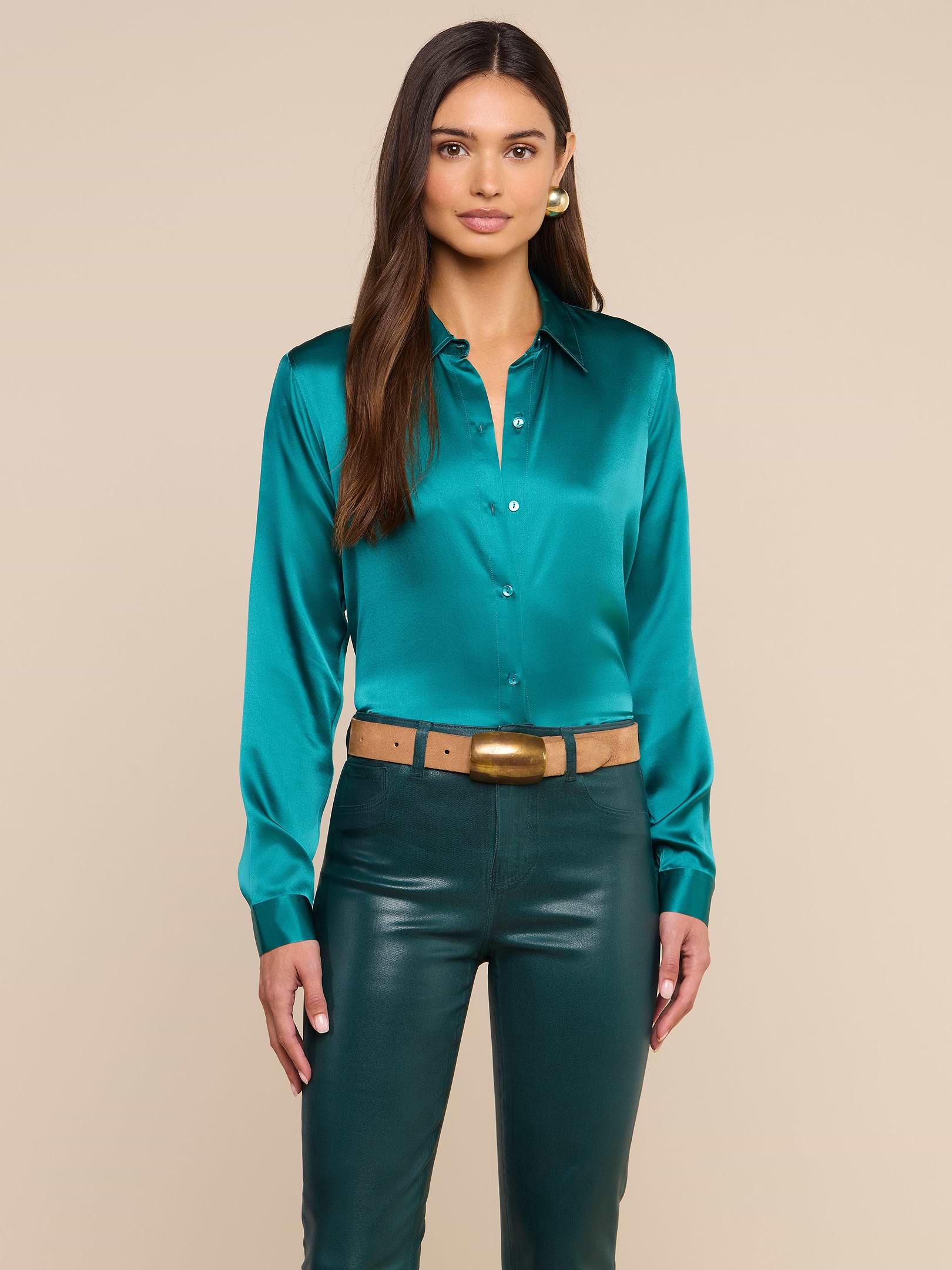 Una donna è in piedi su uno sfondo beige e indossa la camicetta Tyler Silk color verde acqua, abbinata a pantaloni di pelle verde scuro, una cintura color cuoio con fibbia dorata e orecchini dorati. Ha lunghi capelli castani e un’espressione neutra.