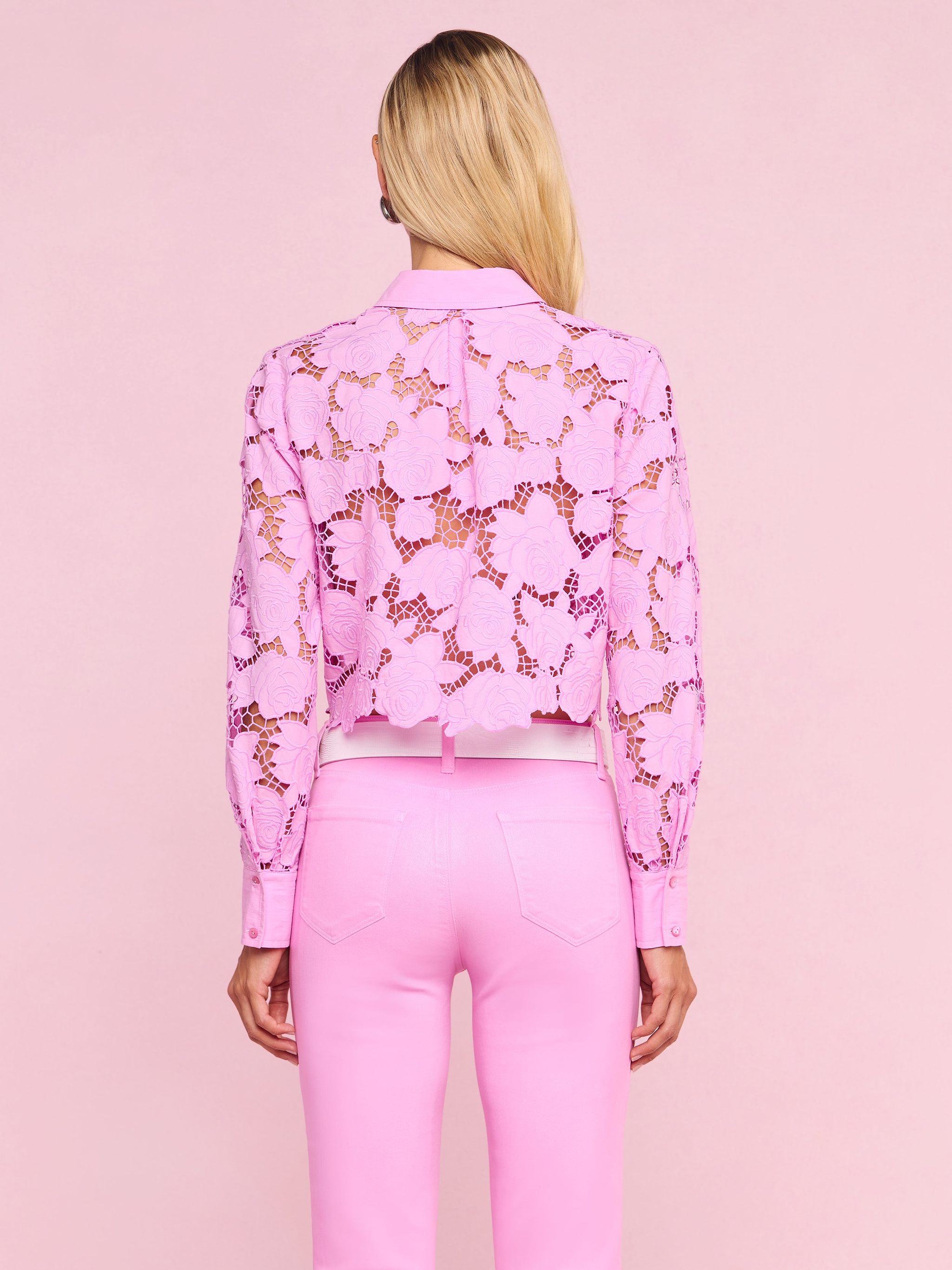A woman stands facing away, wearing light pink pants and the Seychelle Cropped Eyelet Blouse. Her straight blonde hair is down, and the background is plain white.
