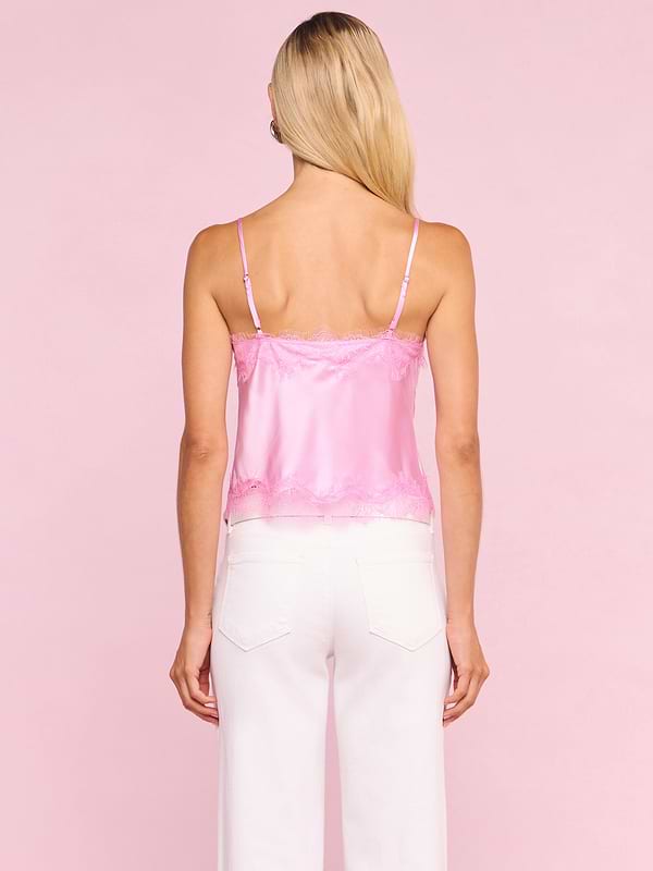 A woman with long blonde hair, facing away, wears the Kimber Silk Lace Camisole from the Legally Blonde capsule collection and white pants against a solid light pink background.