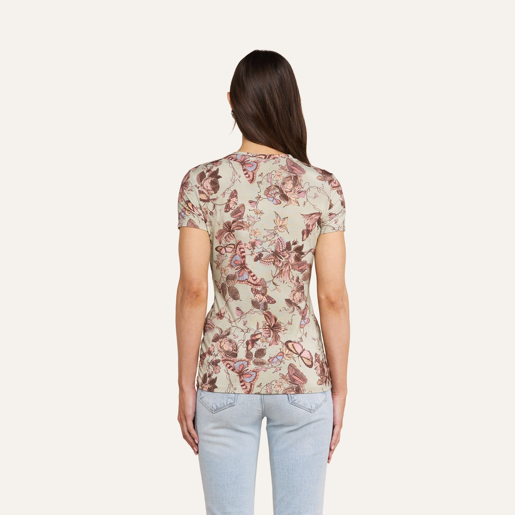 A woman with long brown hair, wearing the beige floral-patterned Ressi Fitted Tee and light blue jeans, stands with her back to the camera against a plain light background.