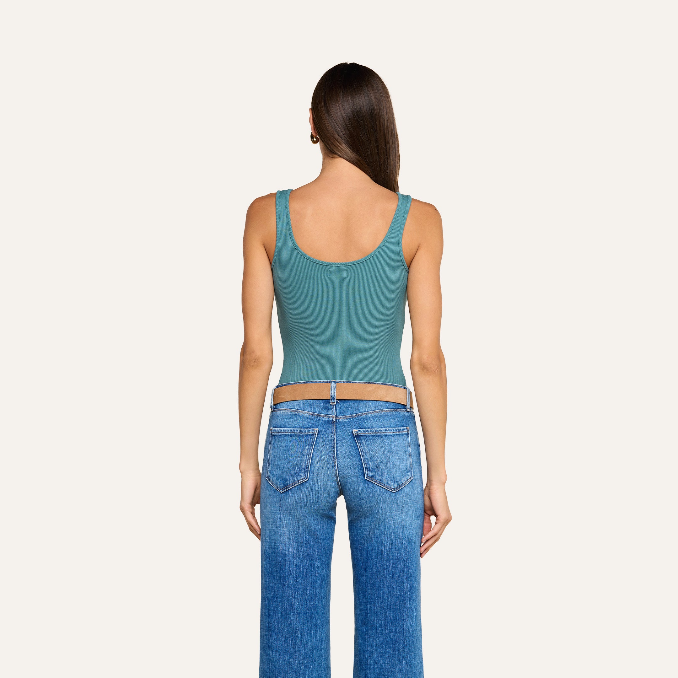 A woman with long brown hair stands facing away, wearing a teal Nica Scoopneck Bodysuit made from high recovery stretch rib and blue jeans. The background is plain and off-white.