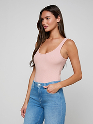A woman with long brown hair wears the Nica Scoopneck Bodysuit in light pink paired with blue jeans, standing with one hand in her pocket and gazing to the side against a plain light background.