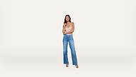 Woman standing against a plain white background, wearing a light pink sleeveless top, blue high-waisted jeans, and heeled sandals, with her arms crossed and looking forward.