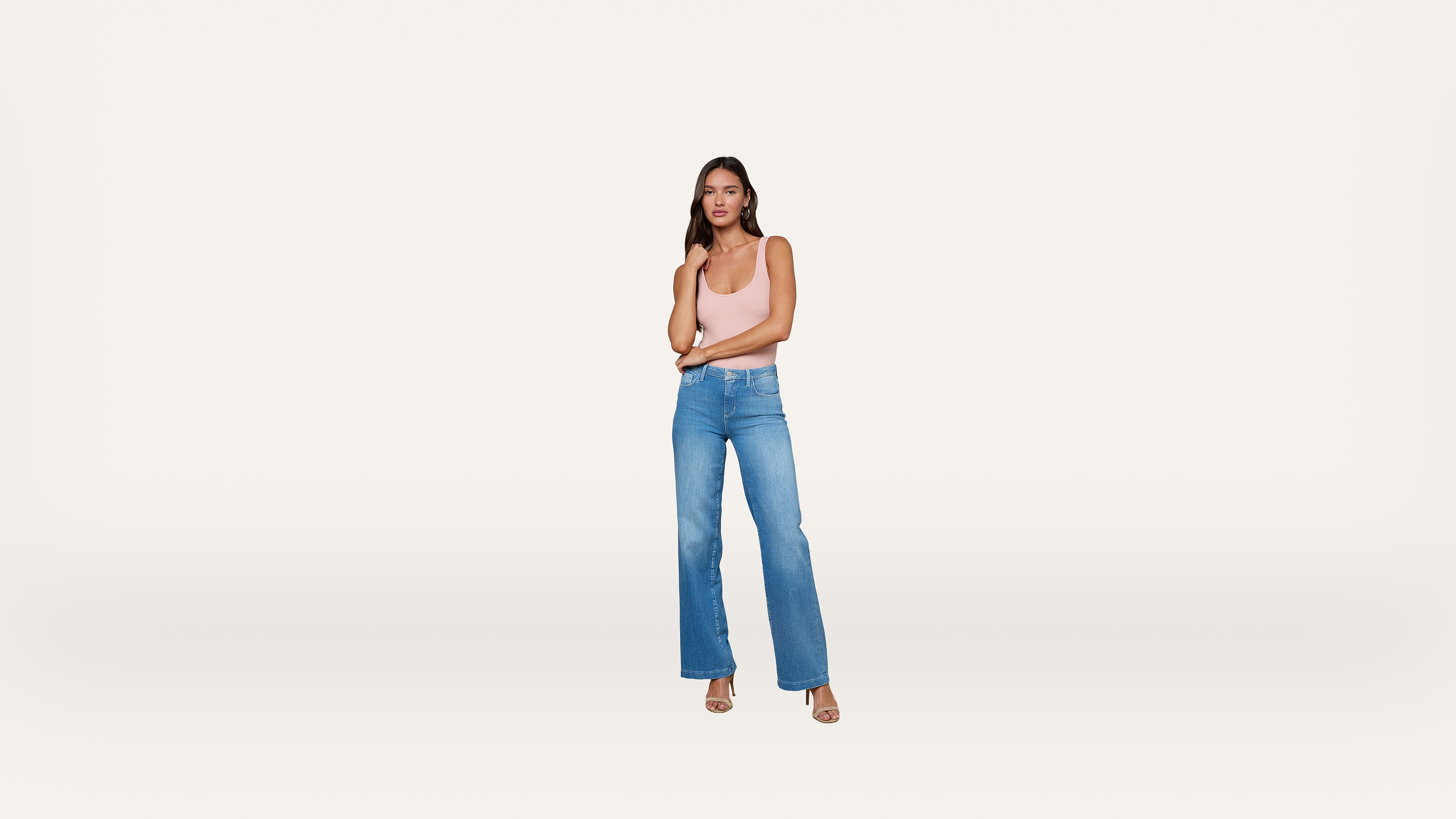 Woman standing against a plain white background, wearing a light pink sleeveless top, blue high-waisted jeans, and heeled sandals, with her arms crossed and looking forward.