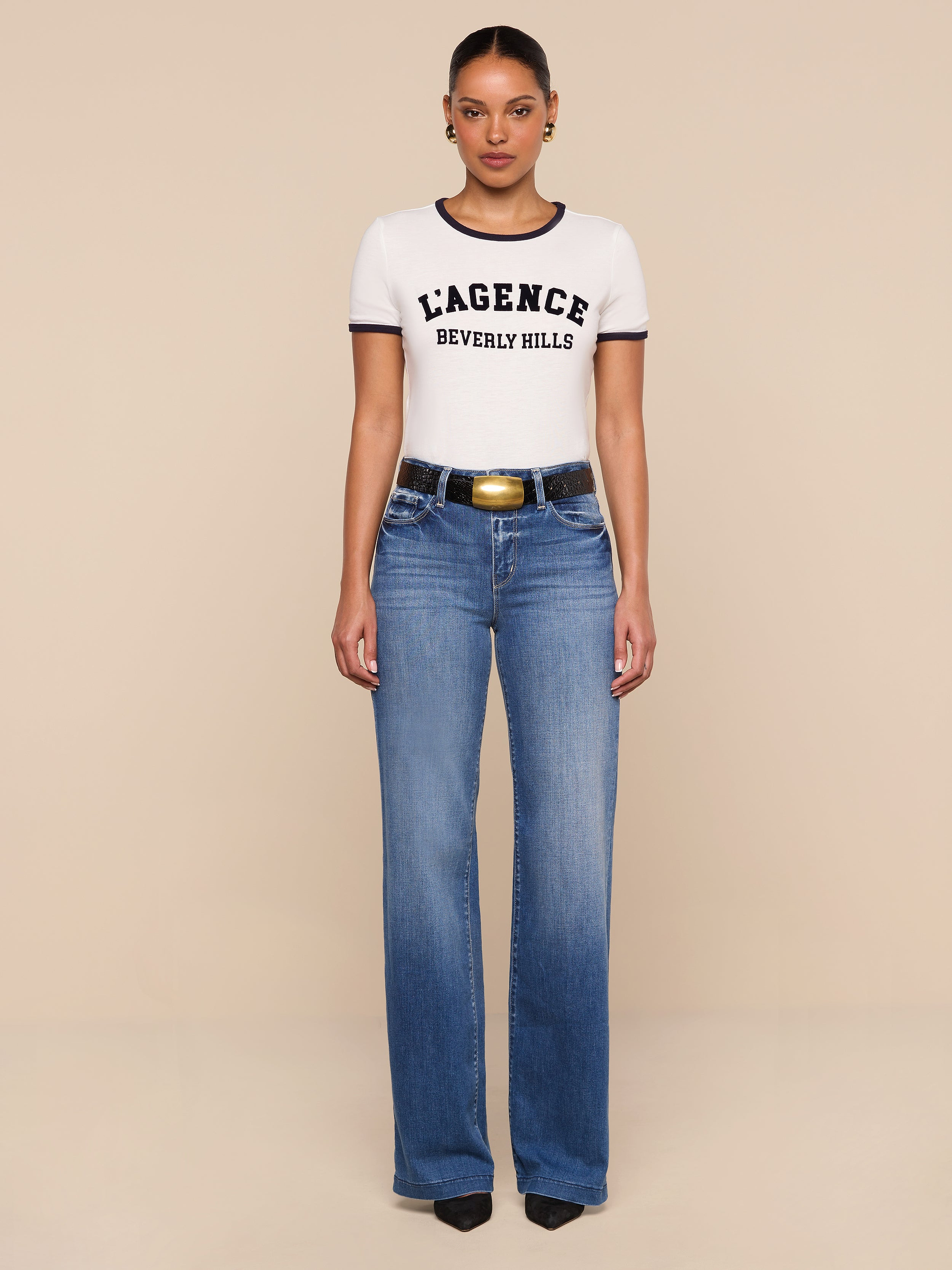 A woman stands facing forward, wearing a relaxed fit white Tommie Crewneck Tee, blue jeans, a black belt with a gold buckle, and black shoes. The background is plain white.