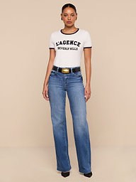 A woman stands against a white background wearing a relaxed fit white Tommie Crewneck Tee made from Supima cotton, blue high-waisted jeans, a black belt with a gold buckle, and black heels.