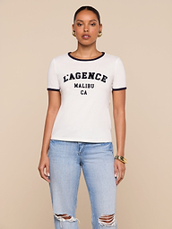 A woman wearing a white Tommie Crewneck Tee with the L’AGENCE logo MALIBU CA text and blue jeans with ripped knees stands facing forward against a plain background.