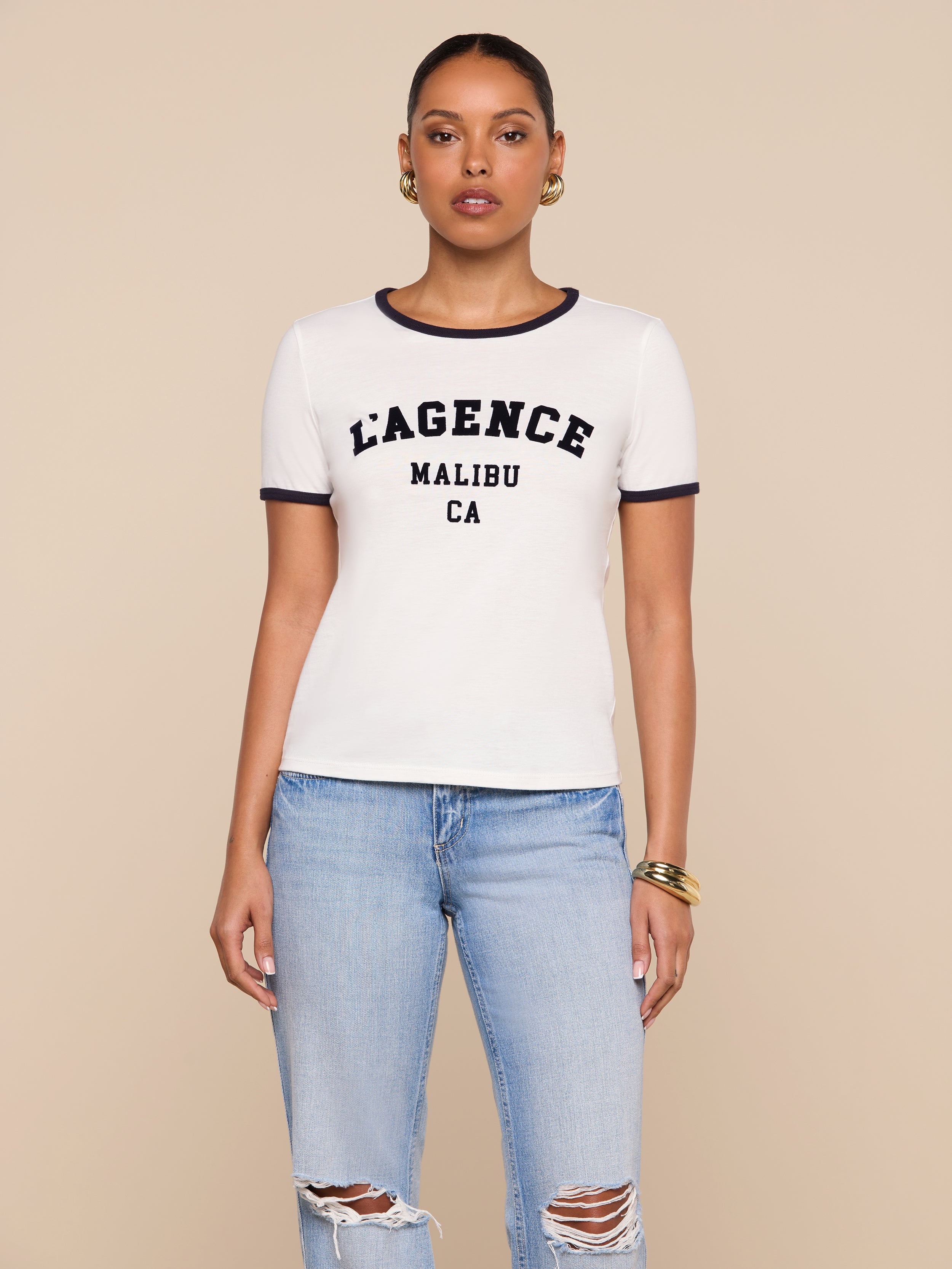A woman wearing a white Tommie Crewneck Tee with the L’AGENCE logo MALIBU CA text and blue jeans with ripped knees stands facing forward against a plain background.
