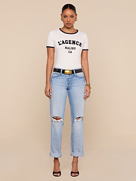 A woman stands against a plain background, wearing a white Tommie Crewneck Tee made from Supima cotton featuring the L’AGENCE logo, light blue ripped jeans, black heeled sandals, a black belt, hoop earrings, and gold bracelets.